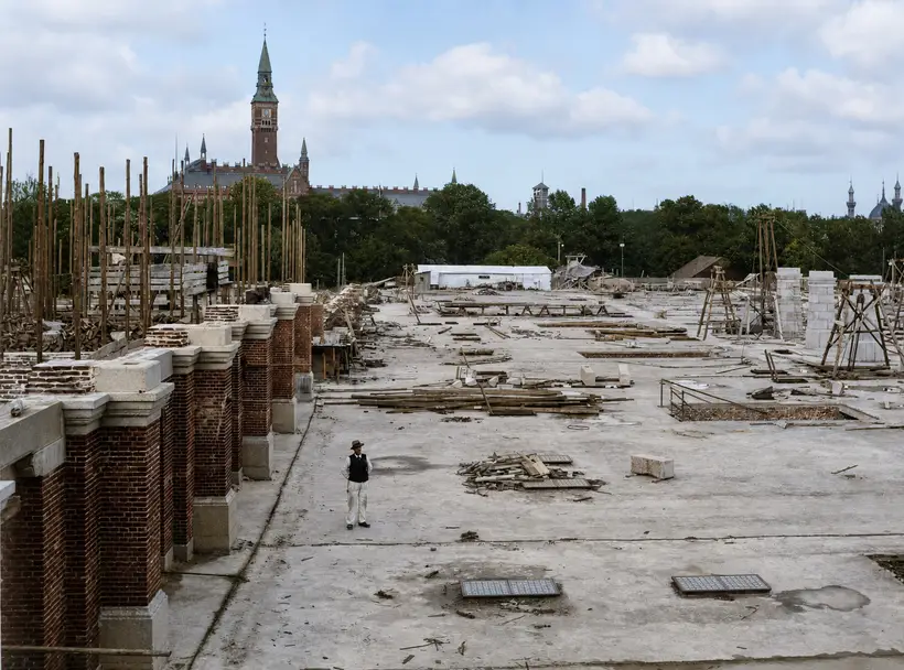 A long open construction deck. At left a row of brick piers with pale granite caps, at right timber trestles and stacks of stone. One man stands alone on the deck, with the tower of Copenhagen City Hall above the treetops.