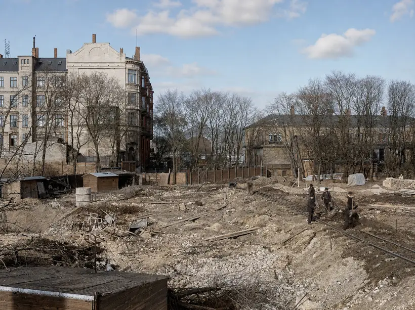 Excavated ground with gravel and rubble and a works track along the floor of the cut. Four men in coats and bowler hats by the track, timber huts at left, a corner apartment block and leafless trees behind.