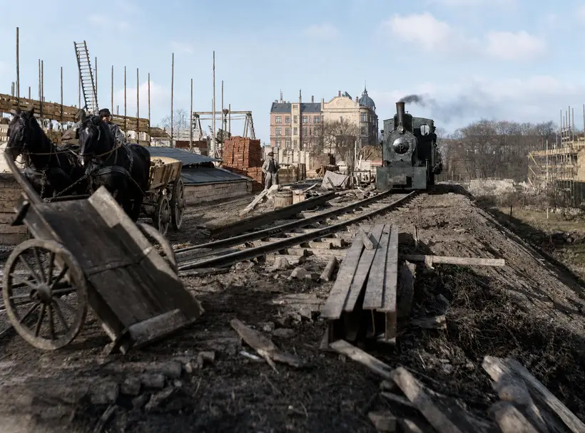 A small steam locomotive under steam on a works track along an embankment. A horse team and cart at left, a tipped handcart in front, stacks of squared timber and red bricks beside the track, bare trees behind.