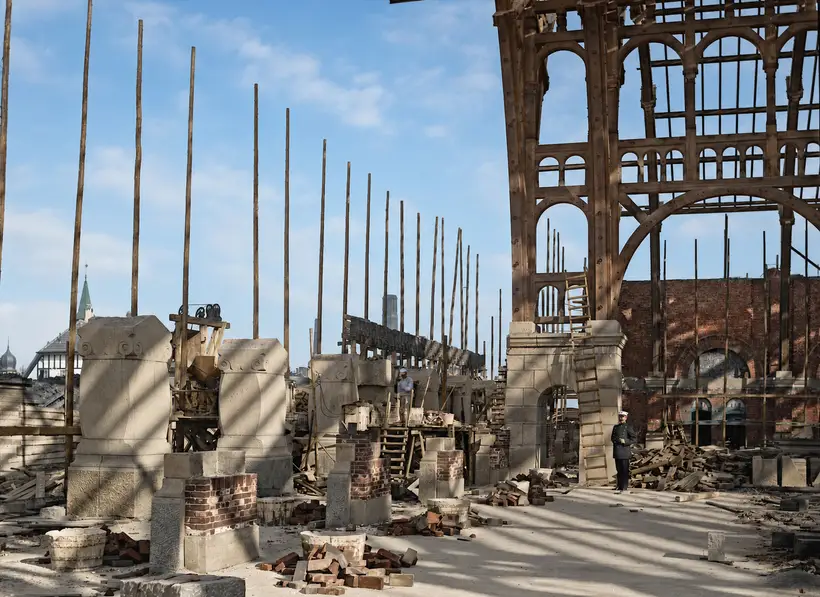 A high-level construction deck: a large arched timber frame with lattice infill on the right, blocks of cut stone in the foreground, and a row of tall thin scaffold poles.