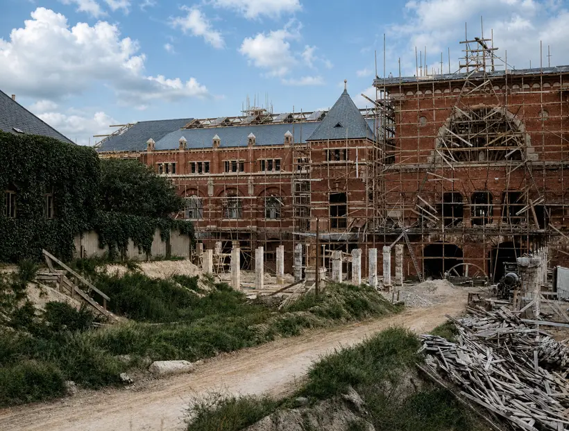 A dirt track through the site, a slated brick wing behind with open roof timbers above the ridge, a heavily scaffolded gable to the right and a large stack of sawn boards in front.