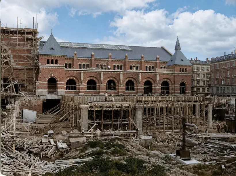A finished brick arcade with a slated roof above a dense mass of timber shoring standing in the track cutting; two stone columns are already up among the timber and workers move across the deck.