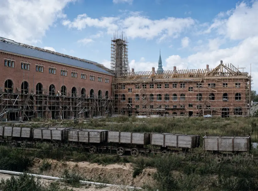 A long brick elevation, half slated and half showing an open timber roof frame, a tall scaffolded shaft in the middle, and a line of open wooden wagons on a narrow track in front.