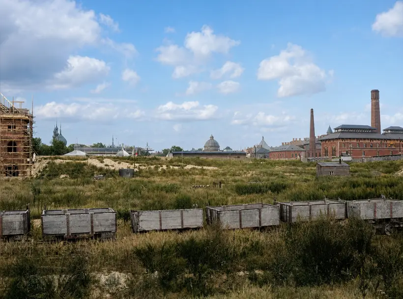 A wide view over cleared, overgrown ground with two lines of open wooden wagons, a hut and sand heaps, a city skyline of domes, spires and chimneys on the horizon, and one scaffolded building corner at the left edge.