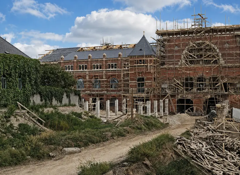A slated brick wing with pale open roof timbers behind it, and to the right a tall gable wrapped in dense scaffolding around a large arch, with a dirt road and stacked boards in front.
