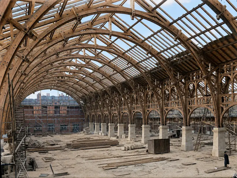 The interior of a large hall roofed entirely in timber — built-up arched ribs, purlins and lattice infill panels — above a sandy floor with free-standing ashlar piers, stacked timber and a single small figure.