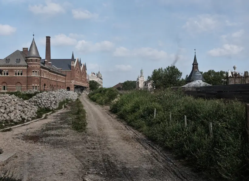A gravel road with deep cart ruts. On the left a long red brick range with a conical corner turret, gables and a tall chimney, and heaps of squared granite along the roadside. On the right scrub, a fence, and Tivoli's spire and glass dome.