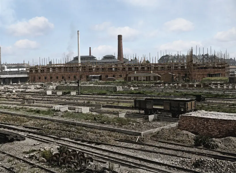 A long red brick building under construction behind a wide railway yard. Round timber scaffold poles rise above the top of the wall along its whole length. Two tall chimneys and factory roofs stand behind it. In the foreground, rails, concrete blocks, a heap of wheelsets, a stack of bricks and an open wooden goods wagon.