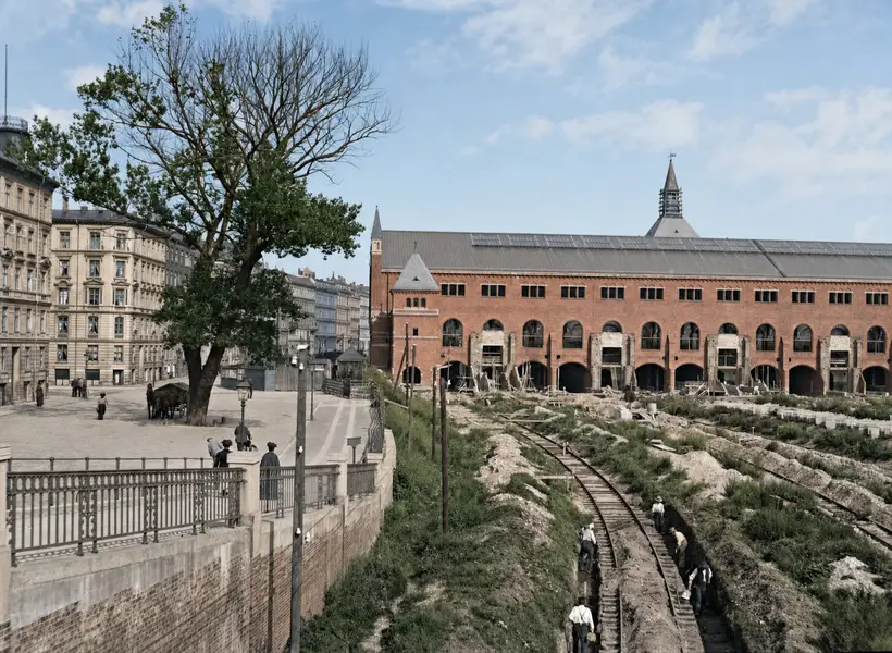 Seen from above: on the left a paved square with an iron railing, trees and tall apartment blocks; on the right a deep excavation where men work along a narrow track. Beyond, a long red brick range with large arches at the bottom under a big slate roof.