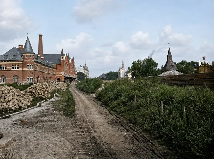 A wide rutted gravel road between two banks. On the left a red brick building with a turret, gables and a chimney, with heaps of granite. On the right stacked timber, a wooden fence, and Tivoli's spire and glass pavilion.
