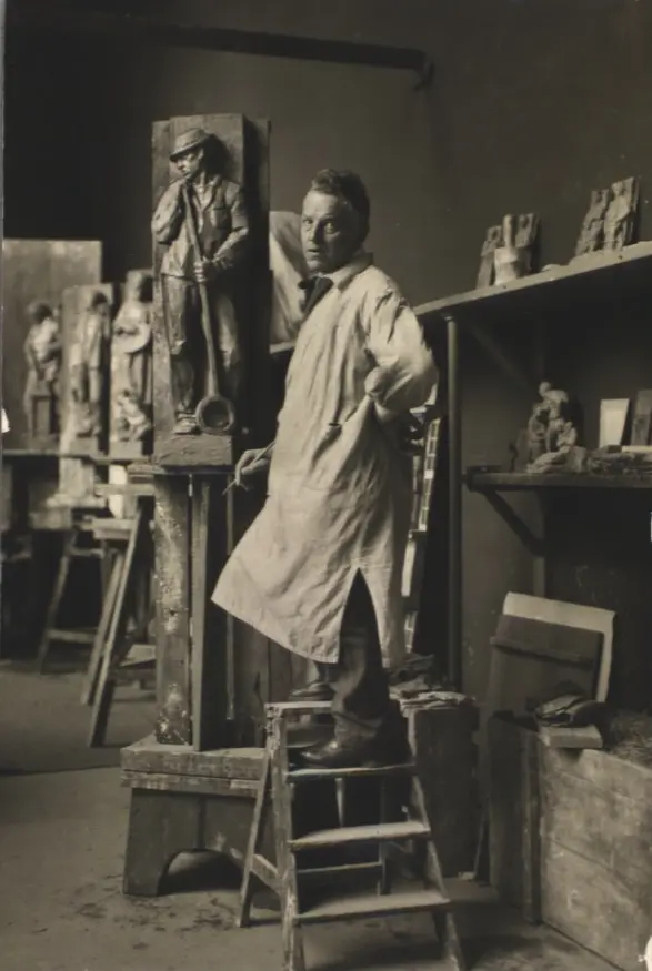 A man in a sculptor's smock stands on a small stepladder with a modelling tool; behind him a clay figure stands against a tall narrow panel.