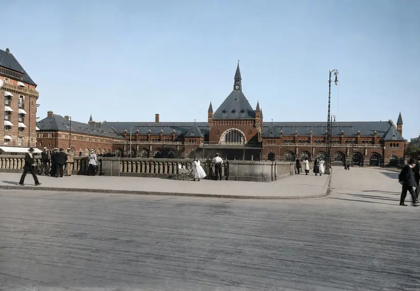 The station's long front seen across a wide open space. At the centre a tall gable with a large round-arched window and a spire above it. In the foreground a stone balustrade with people walking past.