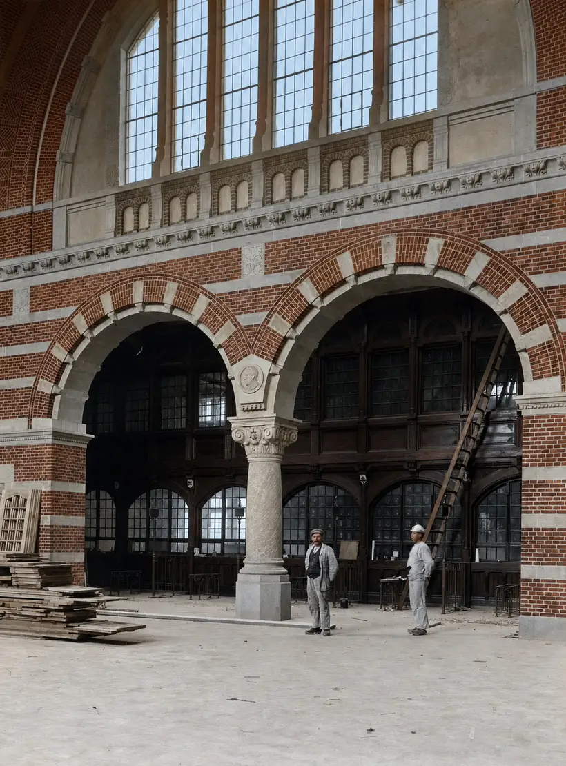 Interior: two large round arches of brick and sandstone carried on a single column with a carved capital. Behind the arches a tall screen of dark joinery with leaded glazing. Two workmen stand on the bare floor; stacked boards lie at the left.