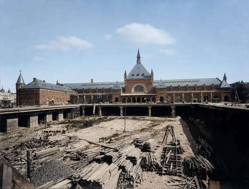 The finished station front seen across a large open excavation. A deck on piers runs across the cutting. The floor of the cutting is covered with stacks of timber, with a timber trestle standing in the middle.