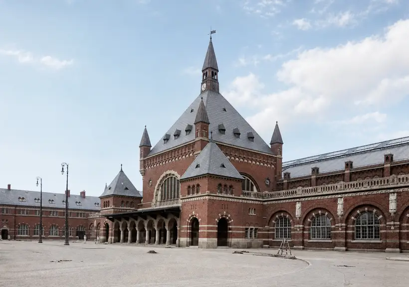 The station's red brick entrance pavilion, with steep slate roof, spire and tall round-arched window, across an empty and unsurfaced forecourt.