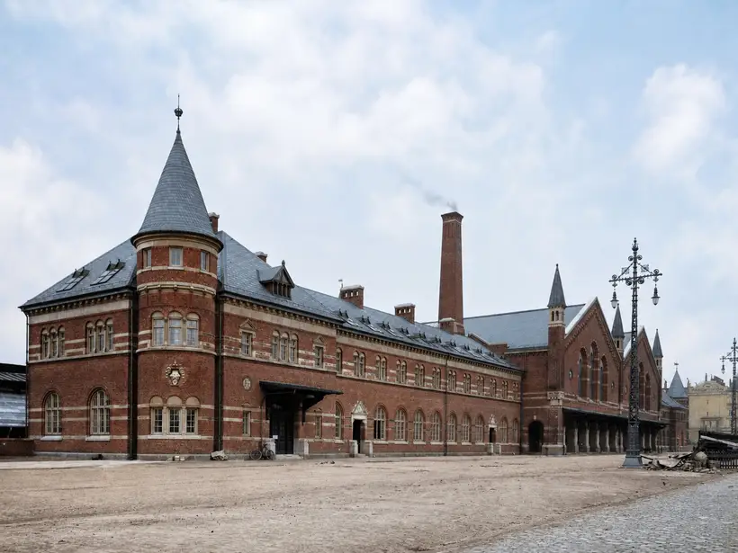 A long red brick wing with round-arched windows, a conical corner tower and a tall chimney, seen along an unmade road.