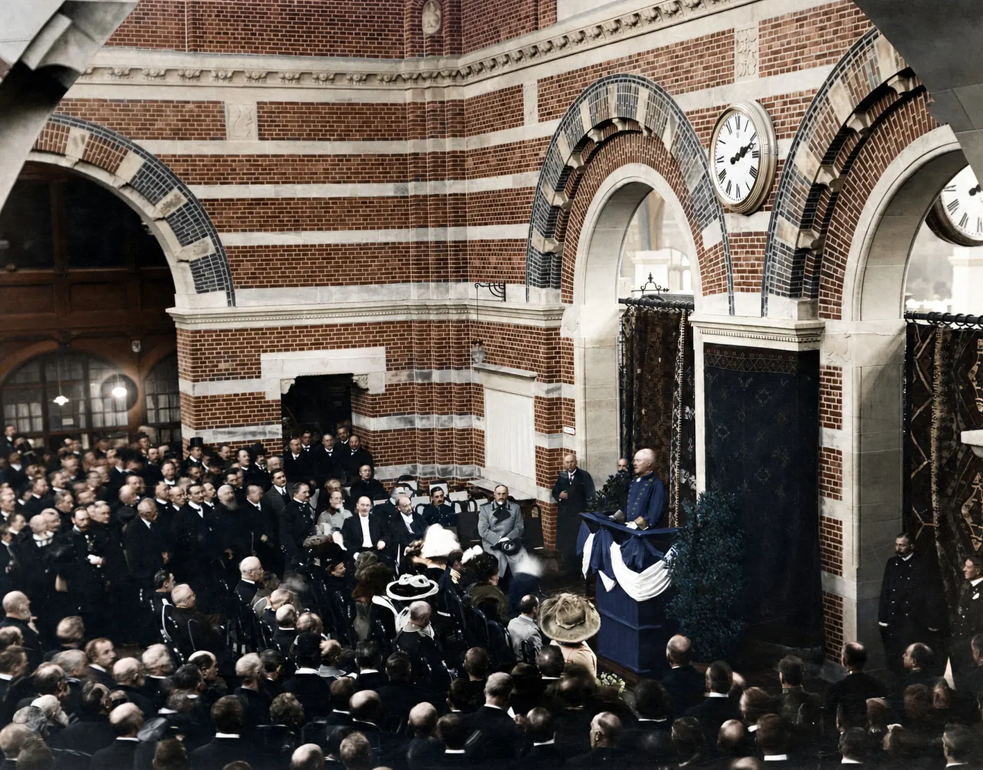A large crowd in coats and hats around a speaker at a draped rostrum, beneath brick arches and a large clock.