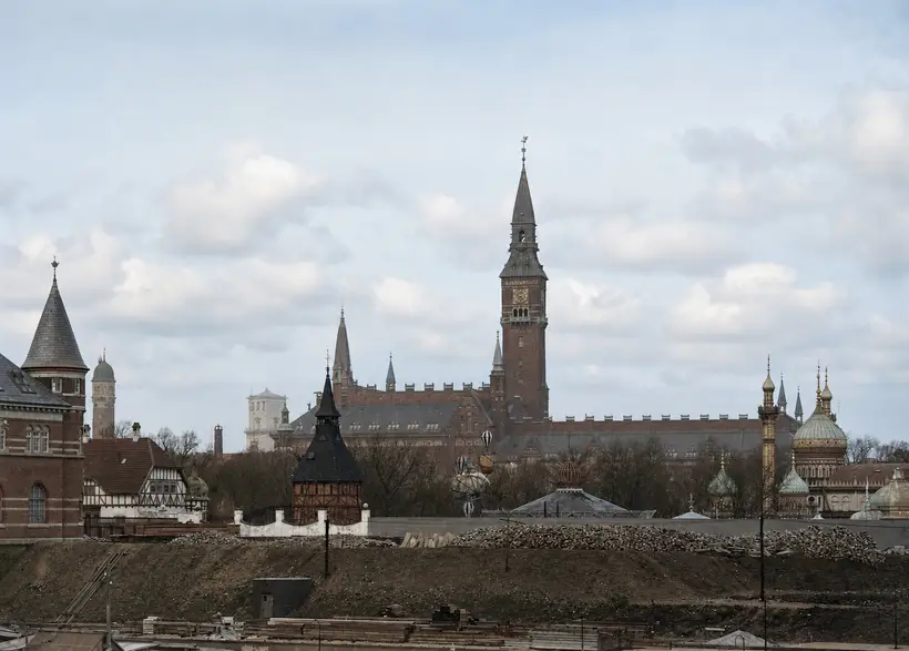 An excavated works site with long piles of timber along the embankment; beyond it a wall, bare trees, the City Hall tower and onion domes.