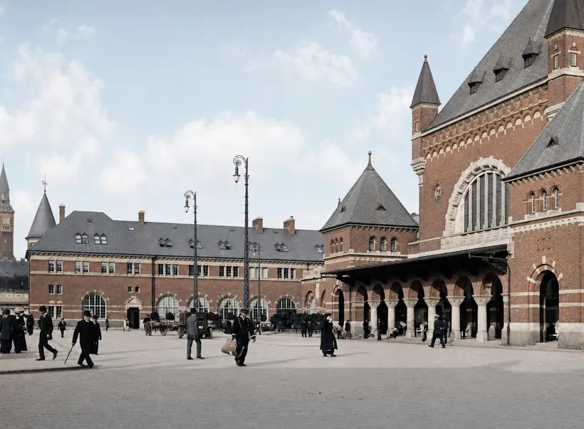 People in long coats and hats crossing a paved forecourt in front of the station's arcade and entrance pavilion.