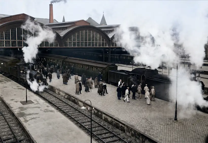 Steam locomotives and waiting passengers on platforms in front of arched glazed gables, under heavy clouds of steam.