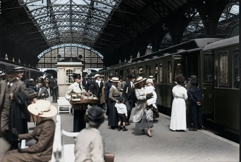 An arched lattice roof structure over a platform with travellers, a kiosk and a green passenger train.