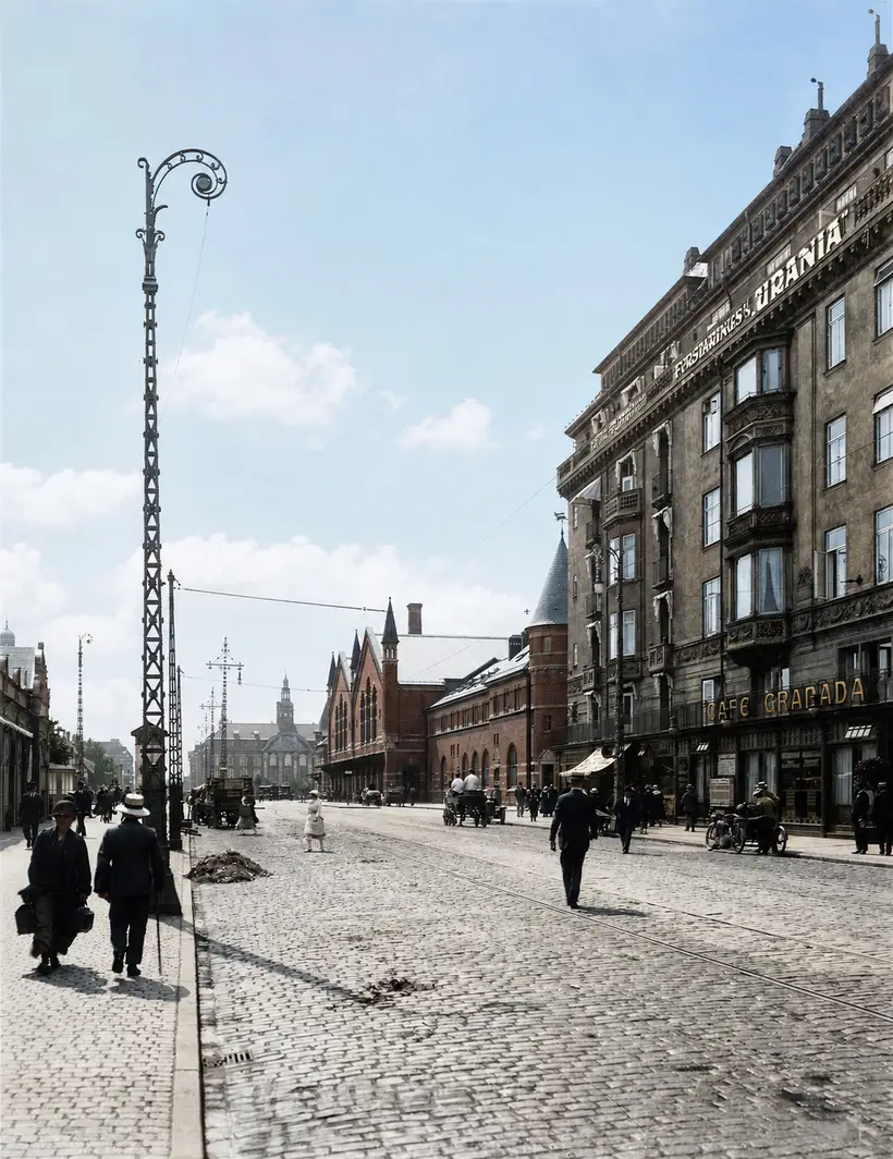 A wide cobbled street with tram rails, a tall lattice mast, pedestrians and a stuccoed block with shop signs on the right.