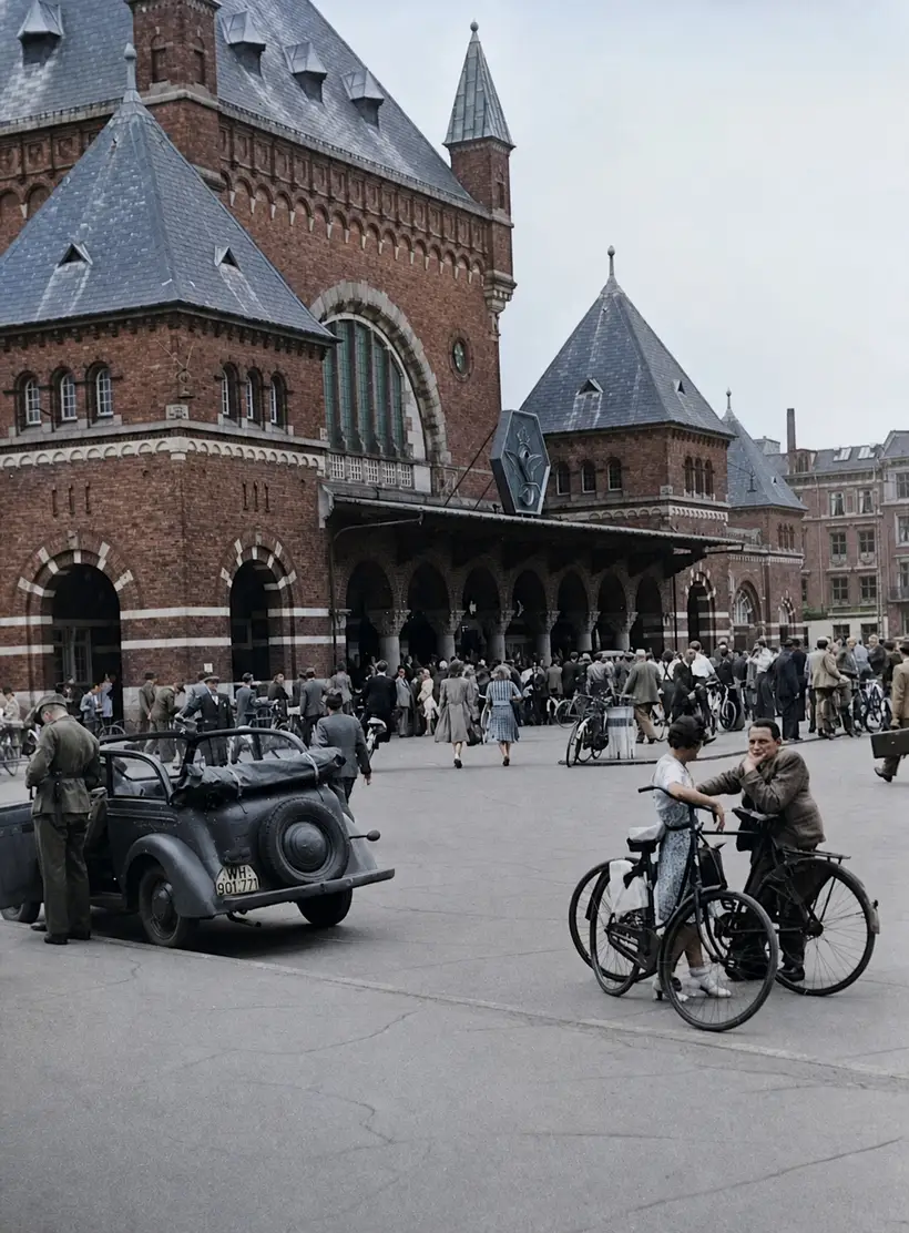 Crowd and bicycles in front of the station's red brick facade with its arcade and slate roofs; an open military car stands at the left.