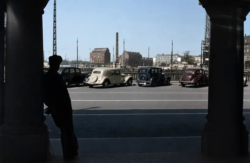 View between two dark columns towards a street with parked 1940s cars, a balustrade, and an obelisk in the distance.