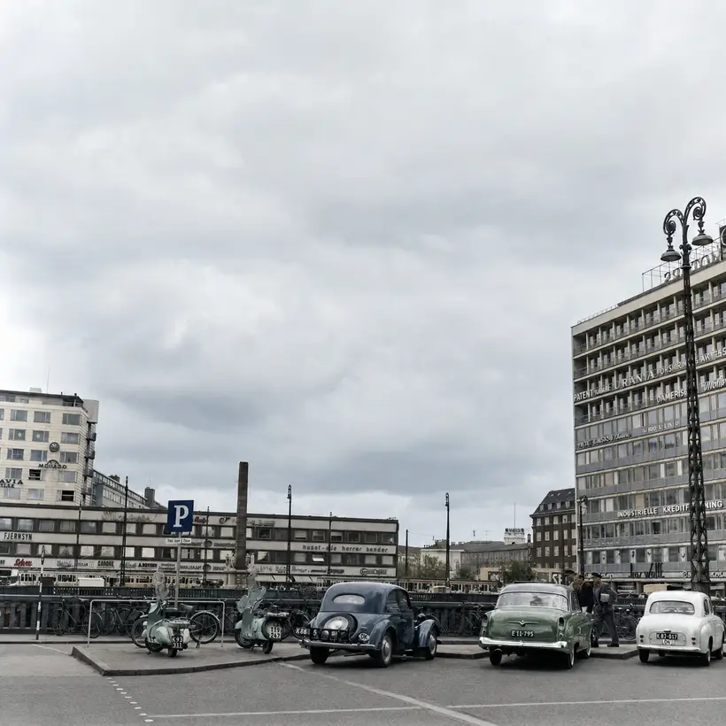 Open forecourt with parked scooters, bicycles and cars in front of tall 1950s office blocks under a grey sky.