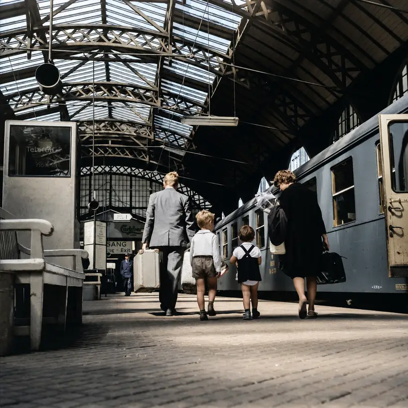 A family with suitcases walks along a platform beneath arched timber roof trusses with a glazed ridge and hanging lamps, beside a blue train.