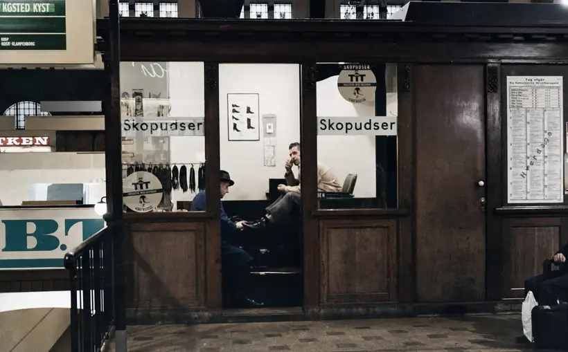 Dark carved timber kiosk lettered Skopudser; inside, a man polishes the shoes of a seated customer.