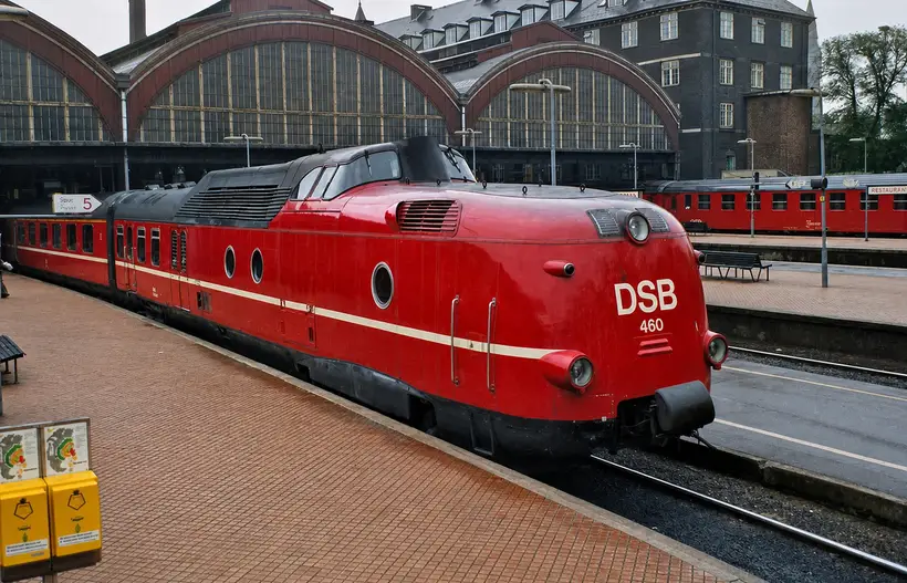 Red streamlined DSB train at a platform in front of three arched glazed gables of the platform halls.