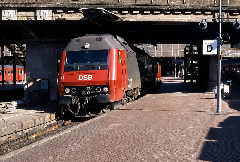 Red and grey DSB locomotive standing in shadow under a road bridge over the tracks, with a paved platform and stairs down from bridge level.