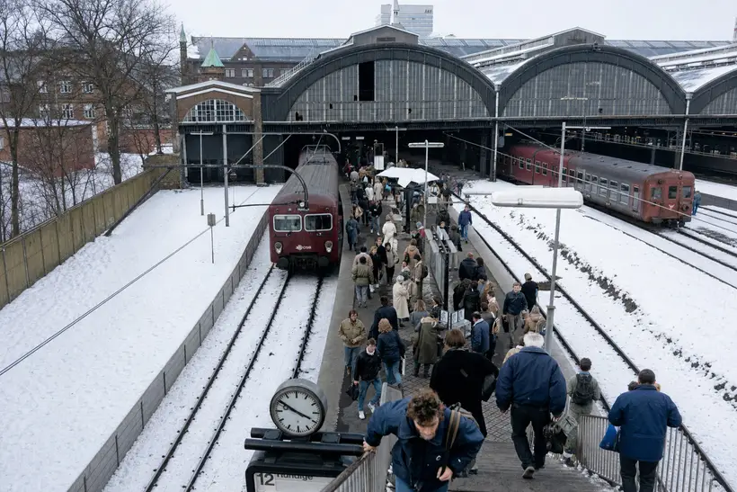 Elevated winter view over snow-covered tracks with two large arched platform halls, red trains, and crowds of travellers on the platform.