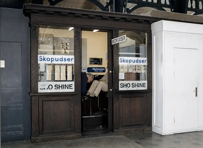 Close view of a carved timber kiosk signed Skopudser and SHO SHINE; inside, a man polishes a seated customer's shoe.
