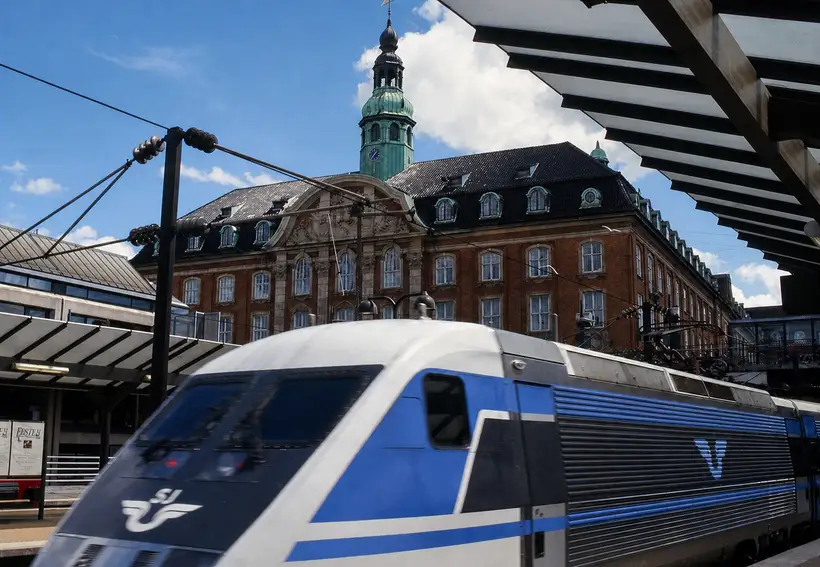 A blue and white electric train at a platform. Behind it a red brick building with a black roof, dormers and a green copper clock tower.