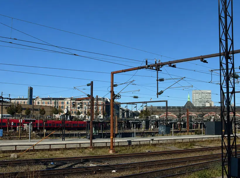 Many tracks and overhead line masts under a blue sky. A red train stands at left, and arched shed roofs are visible far off at right.
