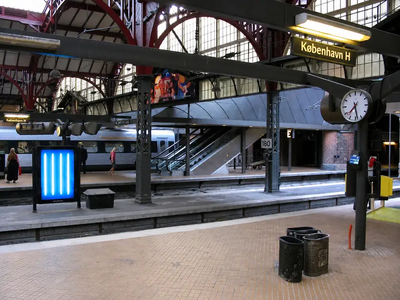 A platform with a Kobenhavn H sign, a clock and escalators. Red arches and high glazing rise above.