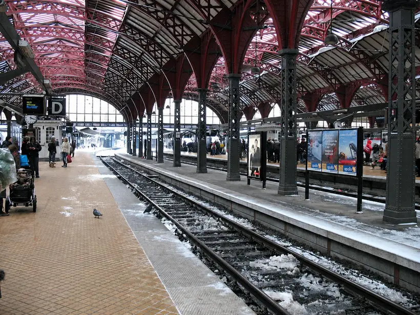 A long platform hall with red arches and lattice bracing overhead. Snow lies between the tracks and people wait on the platform at left.