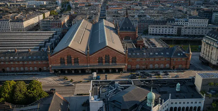 Low aerial of Copenhagen Central Station's entrance front on Bernstorffsgade in warm evening light: two large brick gables with tall round-arched windows and pale stone bands, an arcade behind a projecting canopy at street level, the heavy tower with its steep pyramid roof and tall spire to the north, brick wings running out both ways, and the curved platform hall roofs at the left.