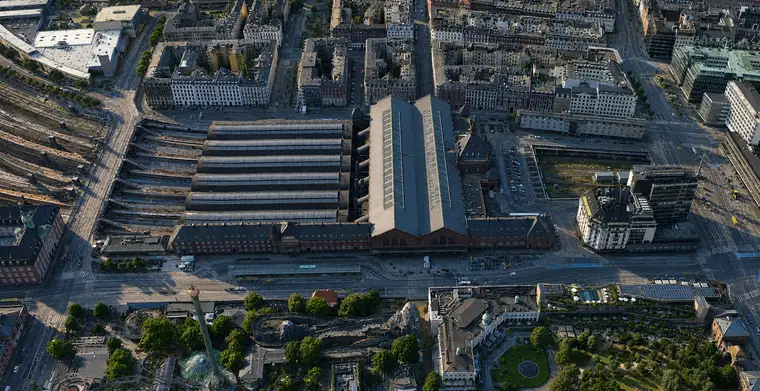 Steep aerial from the east: six long curved glazed hall roofs lie side by side with the great hall closing their head, red brick wings stand between the halls and Bernstorffsgade, and the tracks fan out in the cutting to the south, with Tivoli's gardens along the bottom of the frame.