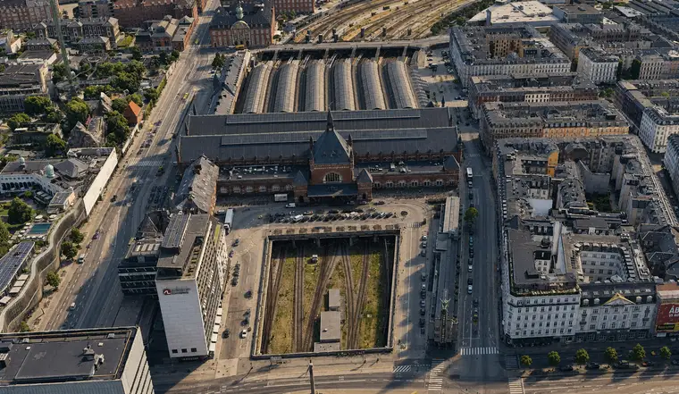 Aerial from north of Copenhagen Central Station: the gabled entrance bay and spire face the camera, six curved hall roofs run away behind, and in the foreground an open cutting carries tracks in under the street through openings in a concrete deck, with five- and six-storey blocks either side.