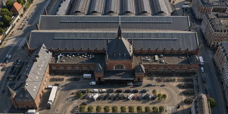 Aerial of the north front of Copenhagen Central Station: red brick with stone dressings, a stepped centre gable carrying a clock and slender spire, long wings either side, the great hall roof behind and the curved platform halls beyond. Cars, trees and ranks of bicycles fill the forecourt below.
