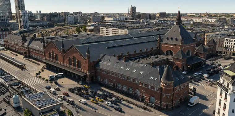 Copenhagen Central Station from the north-east, low above the rooftops. A long red brick range with round-arched windows follows Bernstorffsgade, with taxis and long rows of bicycles below it. At the right the entrance tower and its black spire rise over the great arched window of the main front. Behind lie the glazed roofs of the platform halls, and the tracks fan out into the distance.