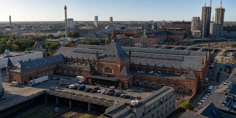 Copenhagen Central Station from the north-west under a clear sky. The main front stands in the middle of the frame with a brick gable, a large arched window and a slender spired tower. A forecourt with parked cars and a lorry lies in front of it. Below that, several tracks run in an open cutting under a road deck carried on concrete piers. To the right the long hall roofs are covered with solar panels. Tivoli's rides and treetops appear at the left, and tower cranes stand on the skyline.