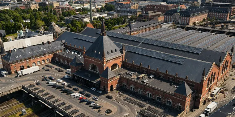 Copenhagen Central Station from the north-west, close and steeply from above. The brick front fills the lower half of the frame, with a tall spired tower, a large arched window and a corner pavilion. Behind it a broad slate roof carries three long strips of solar panels, and beyond that six curved glazed hall roofs run away to the right. Cars and long rows of bicycles stand on the forecourt. Tivoli's trees, green domes and a roller coaster lie at the top left.