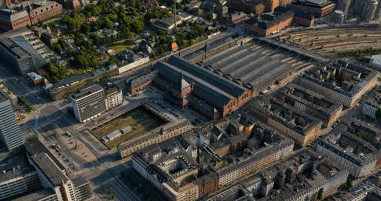 High aerial from the northwest: the great hall crosses the middle of the frame with the spired entrance pavilion on its northern flank, the six platform halls beyond and the tracks fanning out past them. A railway cutting runs toward the camera, Tivoli lies at the upper left and dense nineteenth-century blocks fill the lower half.