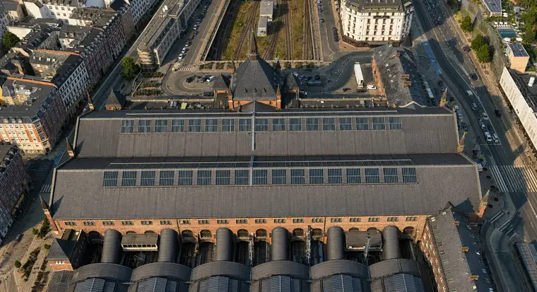 Near-vertical aerial of the great hall roof: a broad grey plane with two long rooflight bands and small spired brick turrets at the corners, the north entrance building's steep roofs and spire beyond, and along the bottom edge the rounded ends of the six vaults meeting a tall brick wall pierced by round-arched openings.