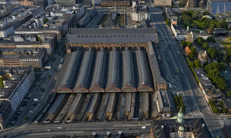 Aerial from the south: six semicircular glazed gables with red-painted arches face the camera, the six glazing-banded barrel vaults run north behind them to the broad roof of the great hall, and in front the tracks lie sunk between the platforms and run out to the street.