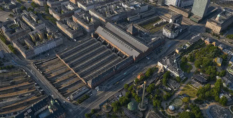 High aerial from the southeast: the six platform halls with their curved gables lie at the lower left, the tracks fan out in an open cutting beyond, the great hall crosses their head with the spired tower beside it, and a second cutting carries the line north between the buildings, with Bernstorffsgade along the front and Tivoli opposite.